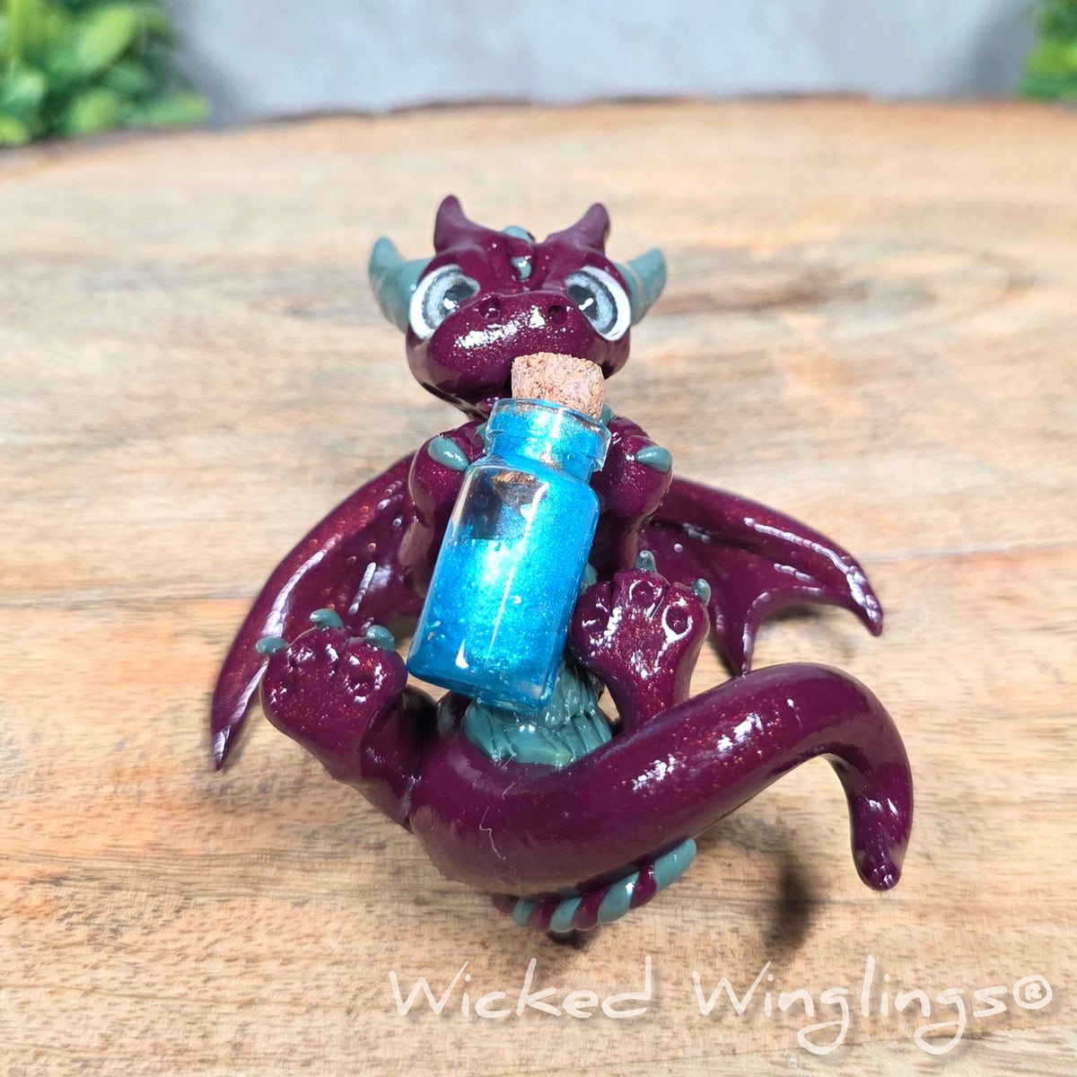 A clay dragon with a potion bottle on a wood surface. Text reads Wicked Winglings.