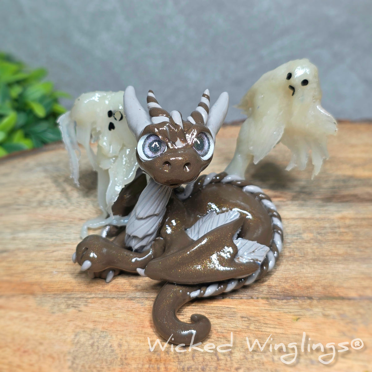 A brown clay dragon with ghosts on a wood surface with greenery. Text reads Wicked Winglings.