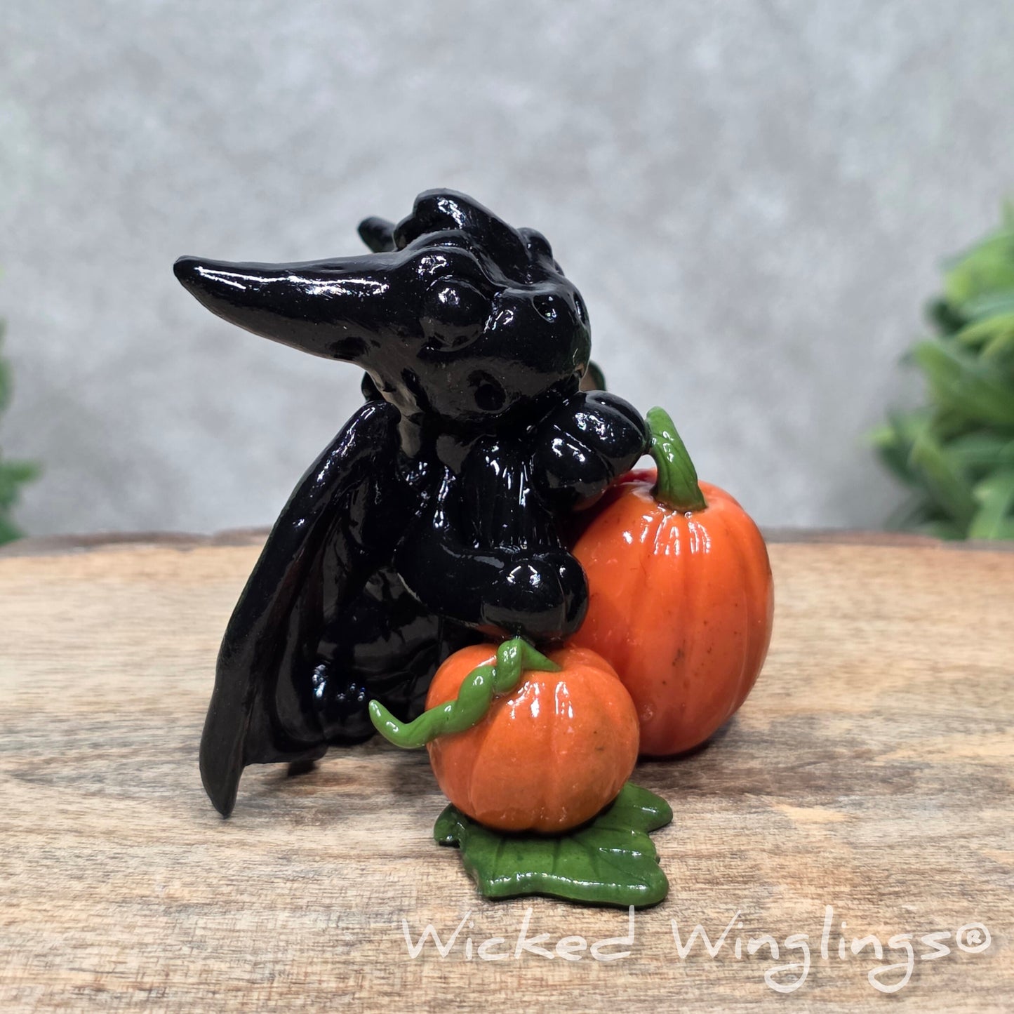 A baby clay dragon sculpture with pumpkins against a wood background with greenery and text Wicked Winglings