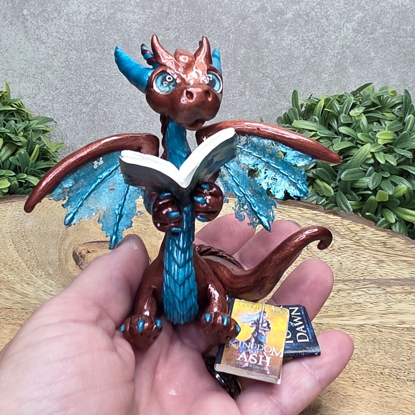 Hand holding a dragon reading a book against a wood surface with greenery