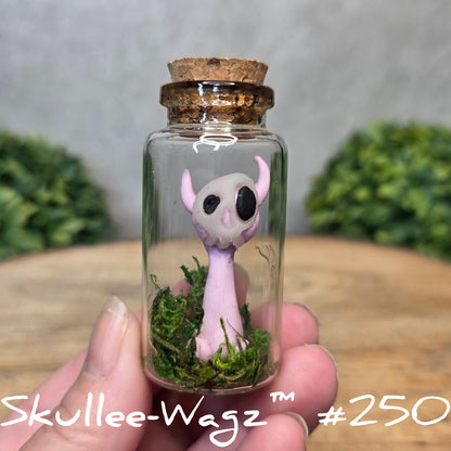 Glass bottle with a skull faced creature and moss, held by a hand with a blurred green background.