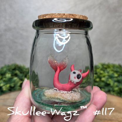 Glass jar with a skull faced creature and sand held by a hand with a blurred green background