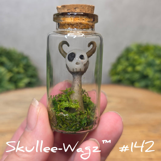 Hand holding a skull faced creature in a jar against a wood background with greenery