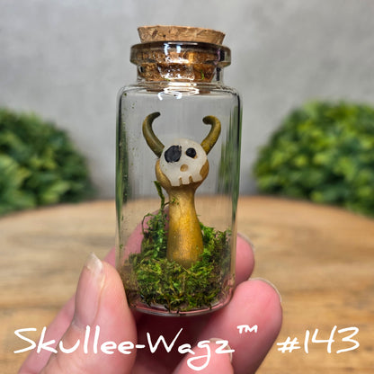 Hand holding a skull faced creature in a jar against a wood background with greenery
