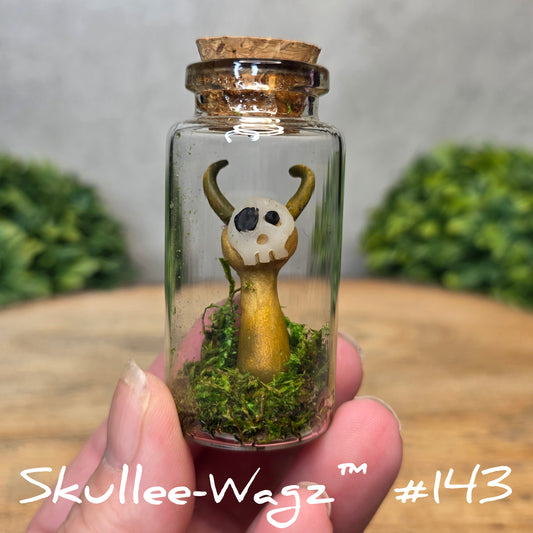 Hand holding a skull faced creature in a jar against a wood background with greenery