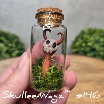 Hand holding a skull faced creature in a jar against a wood background with greenery