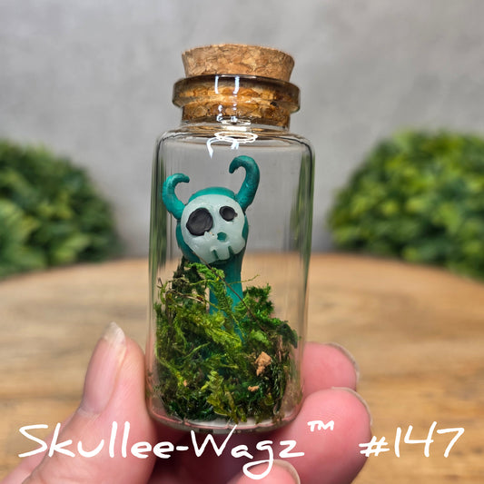 Hand holding a skull faced creature in a jar against a wood background with greenery