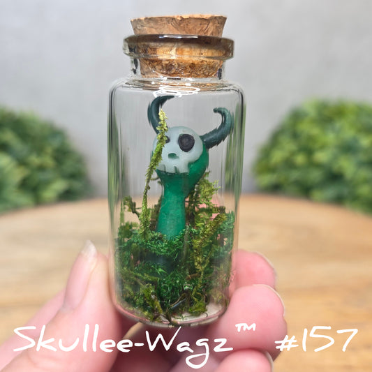Hand holding a skull faced creature in a jar against a wood background with greenery