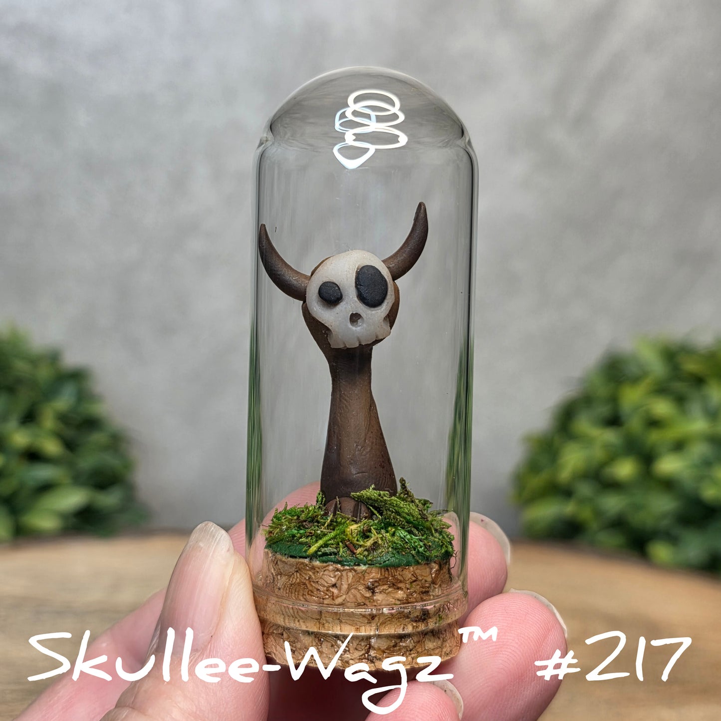 Glass dome with a skull faced creature and moss, held by a hand with a blurred green background.