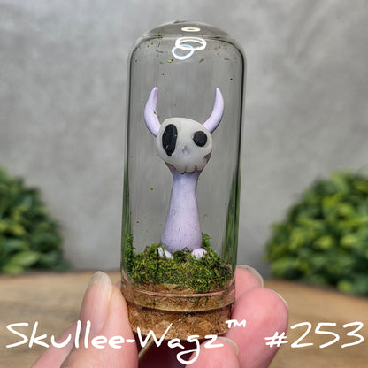 Glass dome with a skull faced creature and moss, held by a hand with a blurred green background.