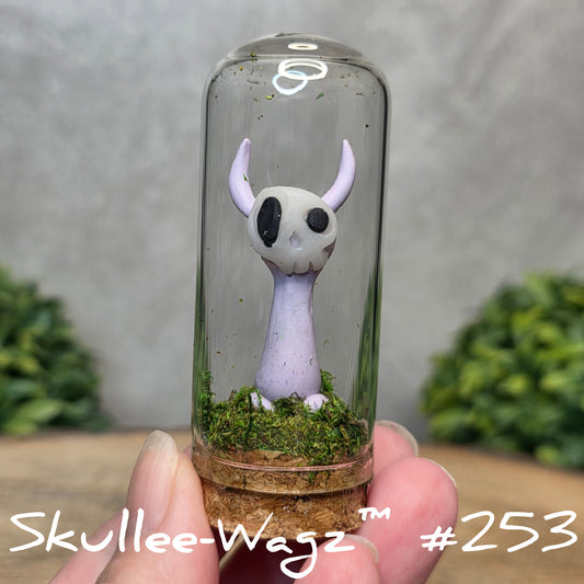 Glass dome with a skull faced creature and moss, held by a hand with a blurred green background.
