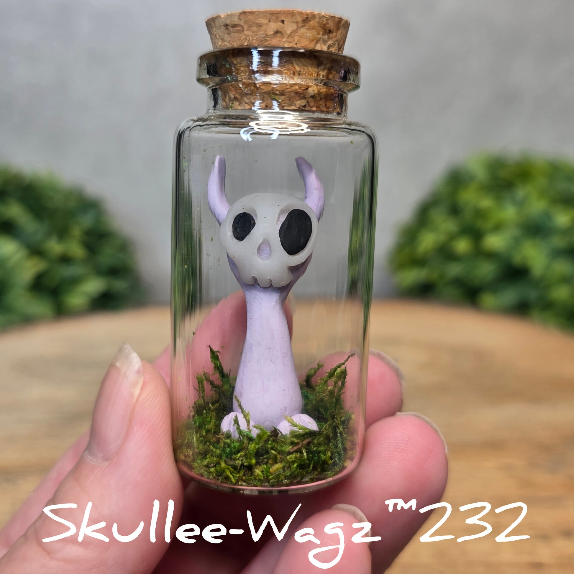Hand holding a skull faced creature in a jar against a wood background with greenery