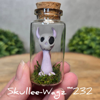 Hand holding a skull faced creature in a jar against a wood background with greenery