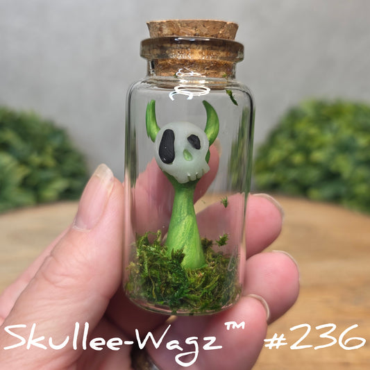 Glass bottle with a skullee-wagz figurine and moss, held by a hand with a blurred green background.