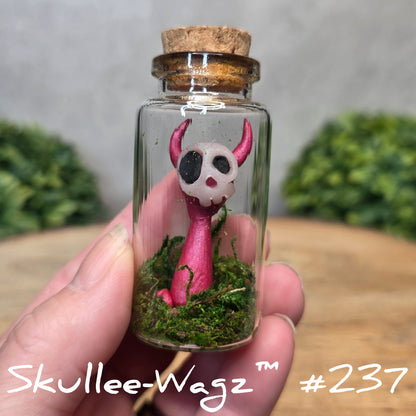 Glass bottle with a skullee-wagz figurine and moss, held by a hand with a blurred green background.