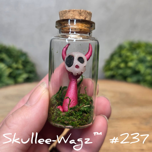 Glass bottle with a skullee-wagz figurine and moss, held by a hand with a blurred green background.