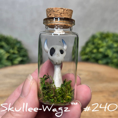 Glass bottle with a skull faced creature and moss, held by a hand with a blurred green background.
