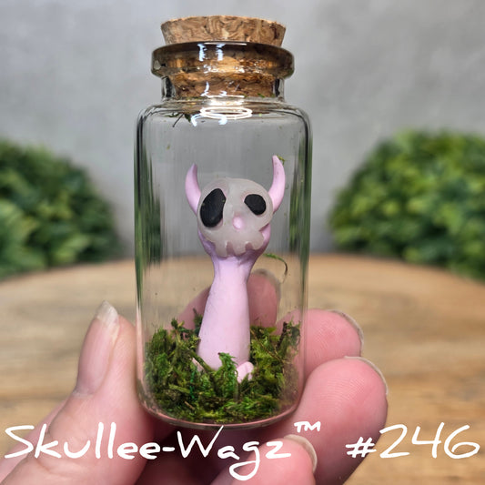 Glass bottle with a skull faced creature and moss, held by a hand with a blurred green background.