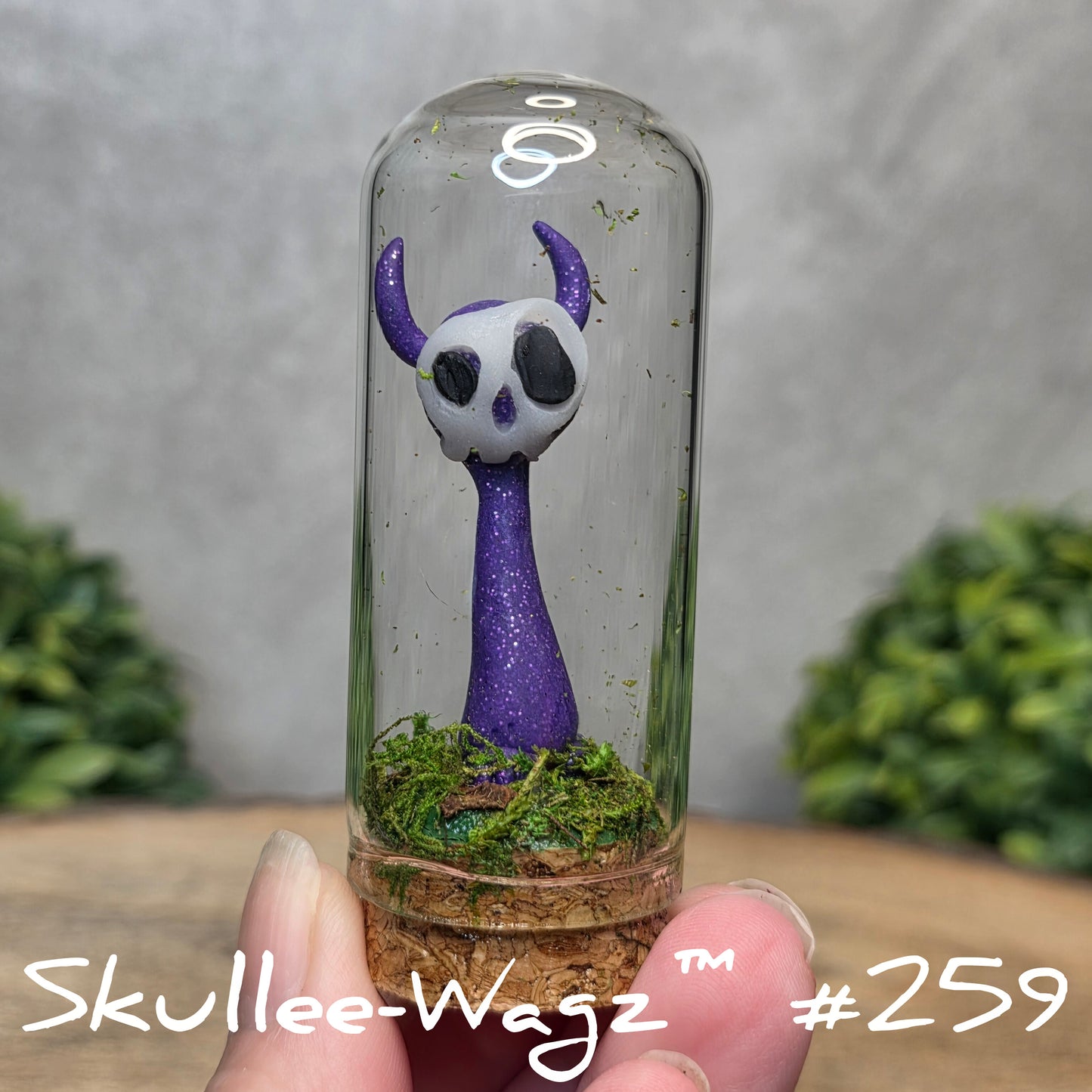 Glass dome with a skull faced creature and moss, held by a hand with a blurred green background.