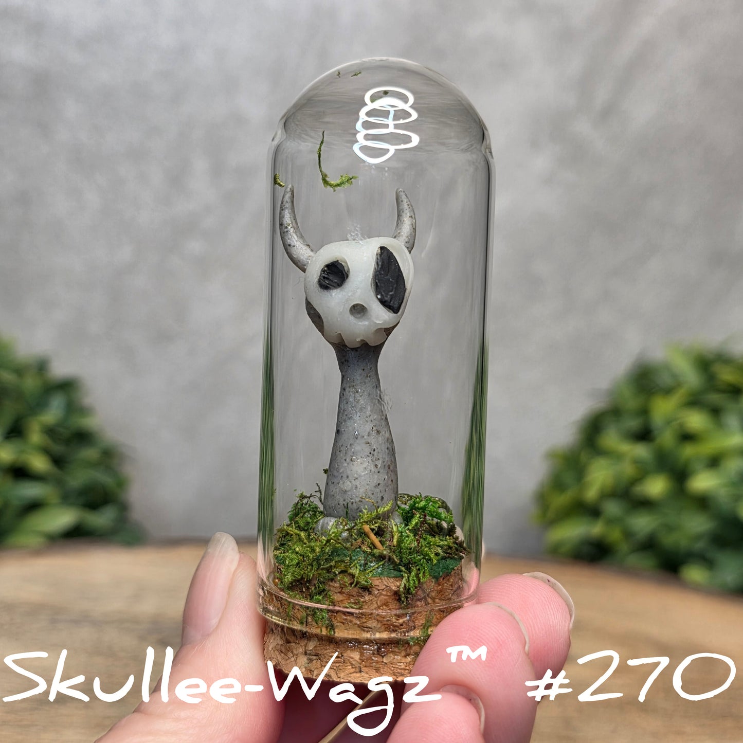 Glass dome with a skull faced creature and moss, held by a hand with a blurred green background.