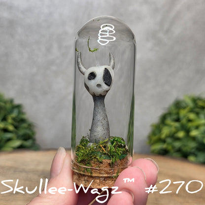 Glass dome with a skull faced creature and moss, held by a hand with a blurred green background.