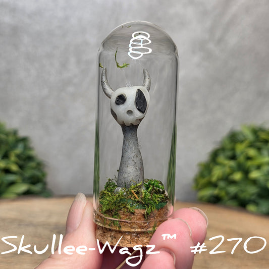 Glass dome with a skull faced creature and moss, held by a hand with a blurred green background.