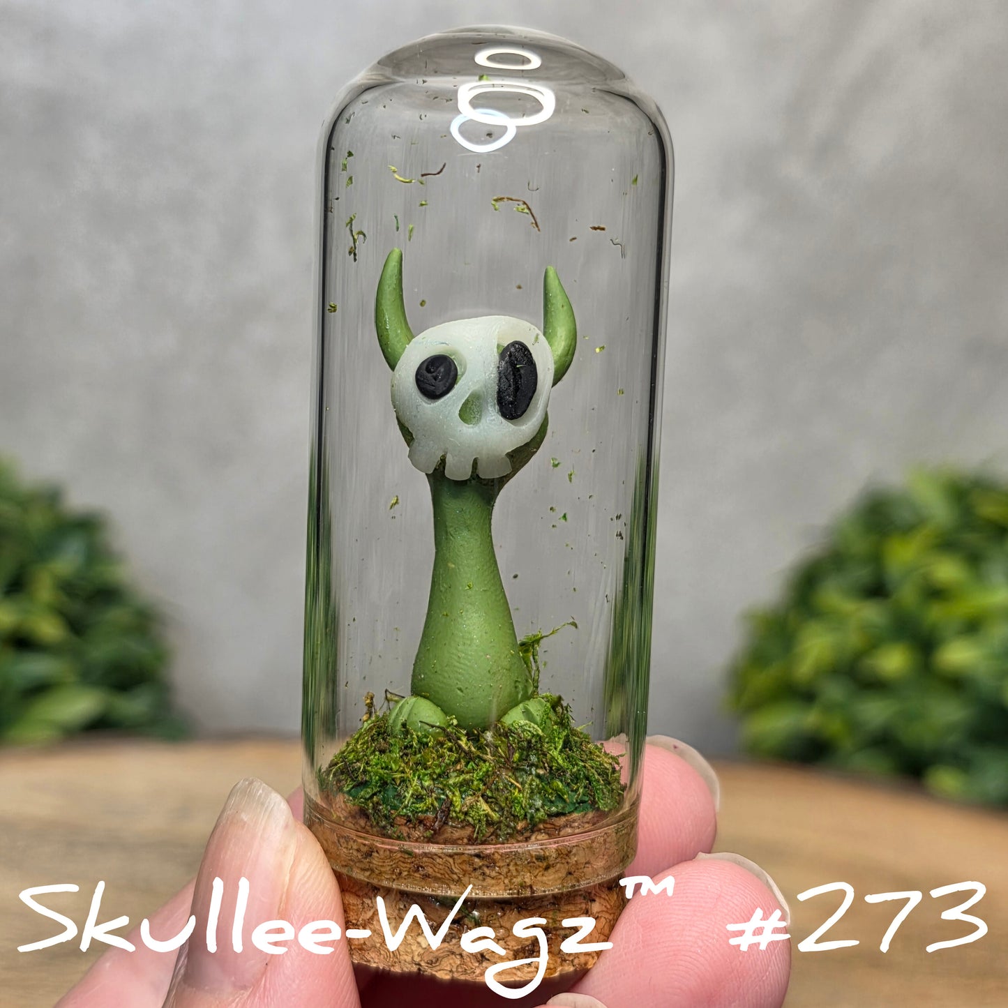 Glass dome with a skull faced creature and moss, held by a hand with a blurred green background.