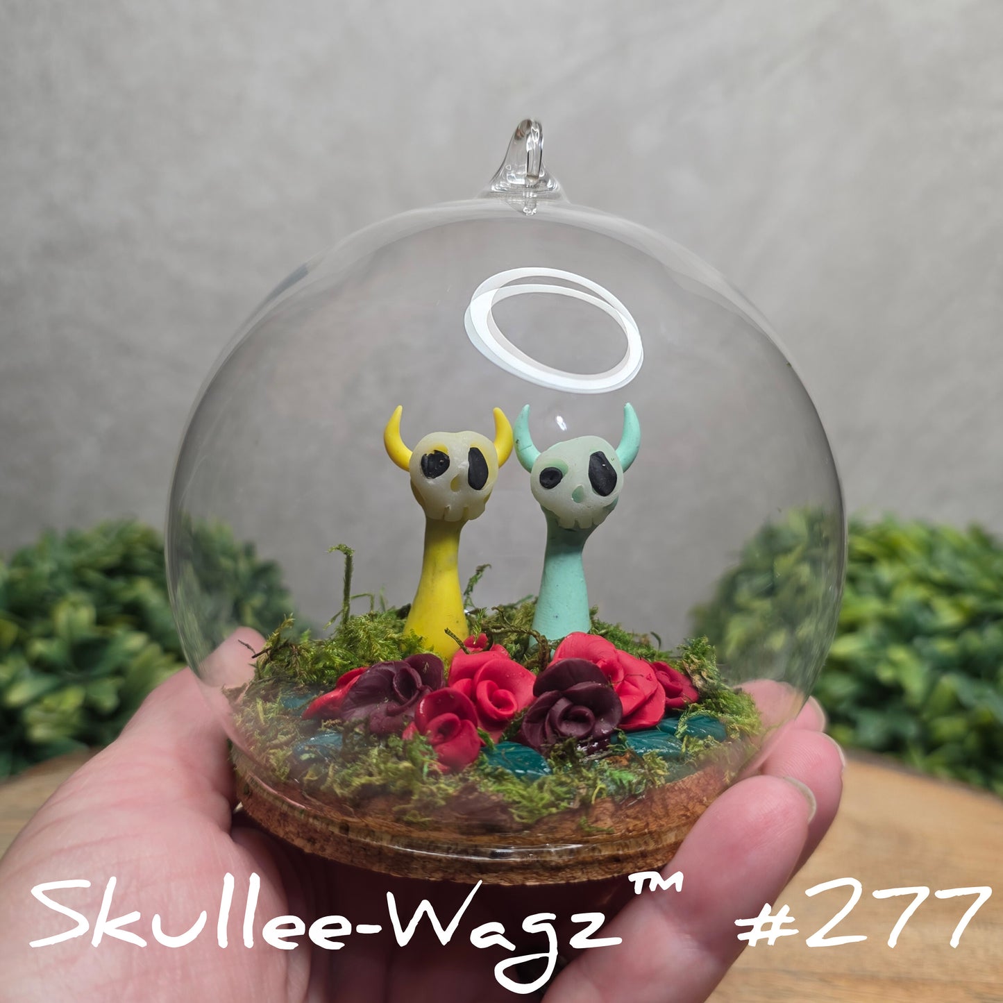 Glass dome with a skull faced creatures, clay roses and moss, held by a hand with a blurred green background.