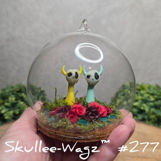 Glass dome with a skull faced creatures, clay roses and moss, held by a hand with a blurred green background.