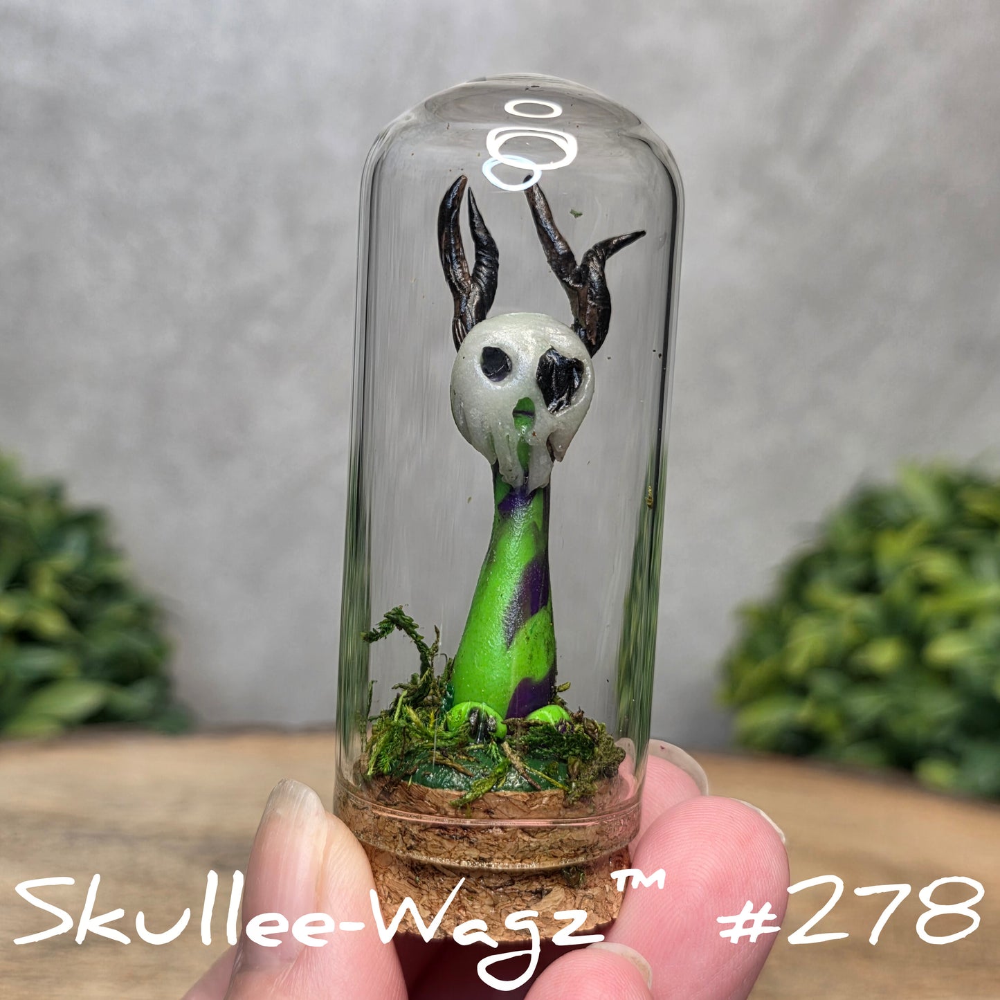 Glass dome with a skull faced creature and moss, held by a hand with a blurred green background.