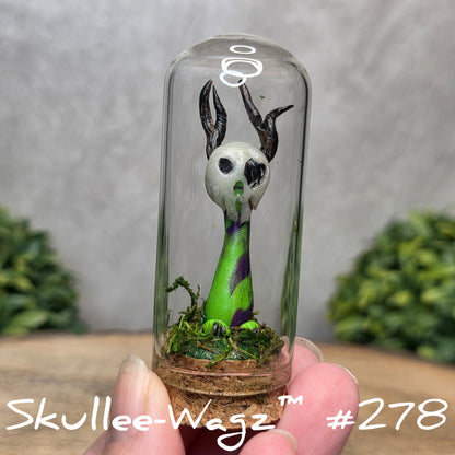 Glass dome with a skull faced creature and moss, held by a hand with a blurred green background.