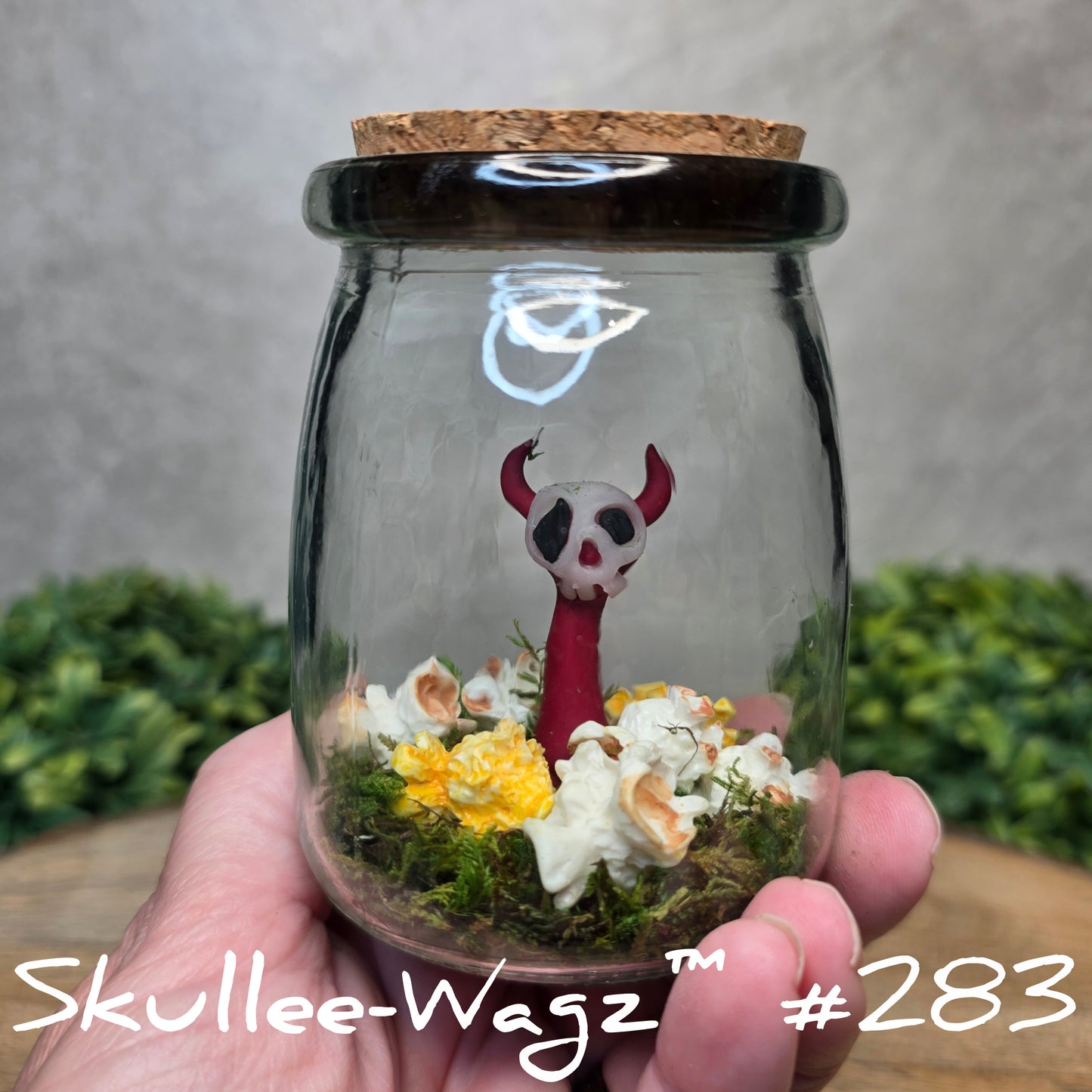 Glass jar with a skull faced creature, resin popcorn and moss, held by a hand with a blurred green background.