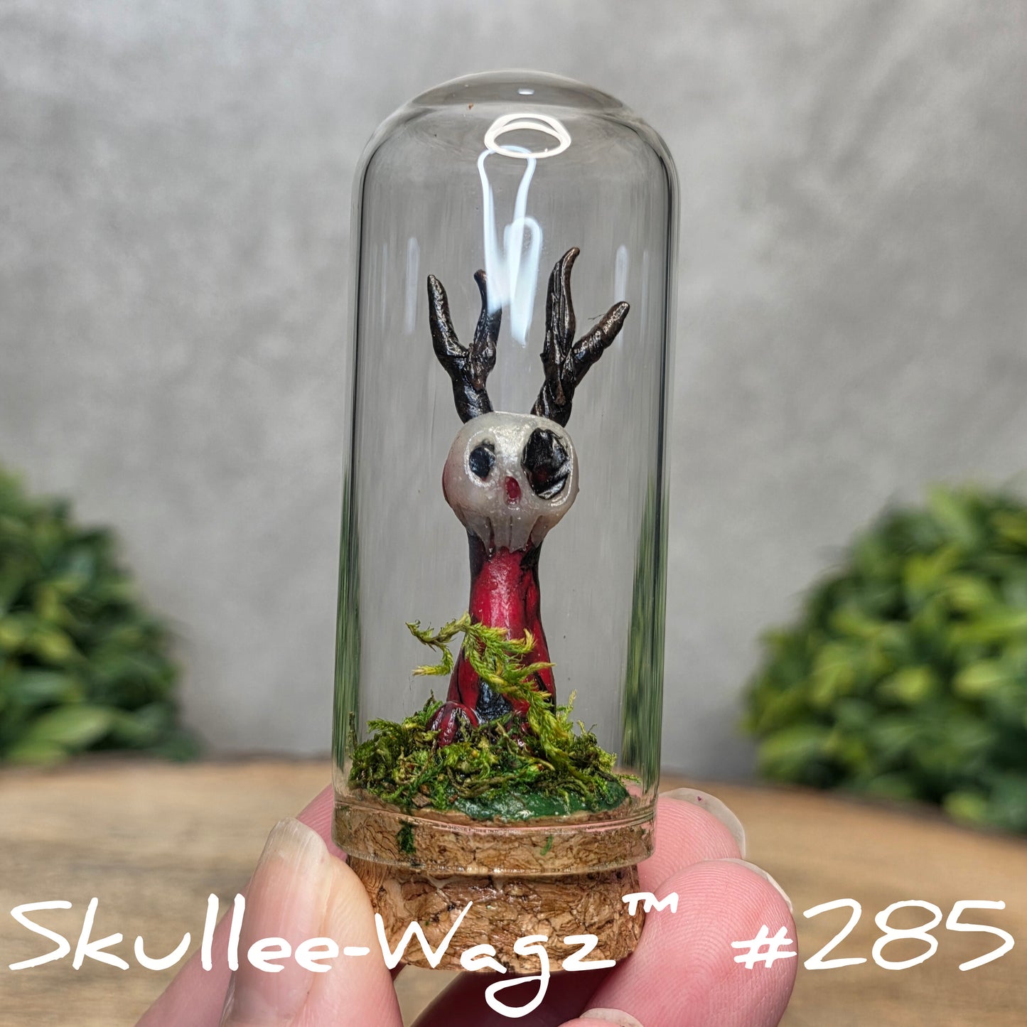 Glass dome with a skull faced creature and moss, held by a hand with a blurred green background.