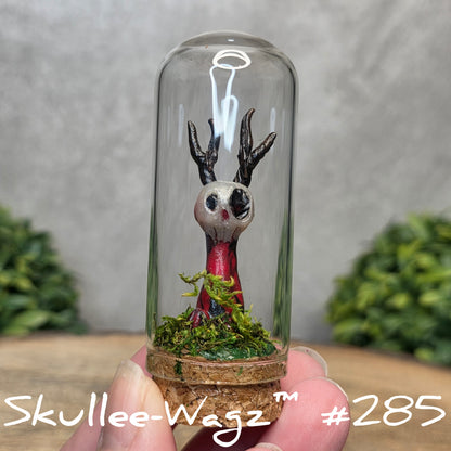 Glass dome with a skull faced creature and moss, held by a hand with a blurred green background.