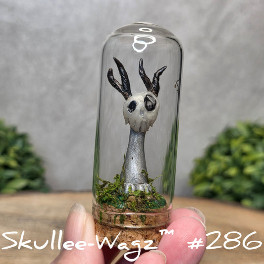 Glass dome with a skull faced creature and moss, held by a hand with a blurred green background.
