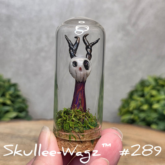 Glass dome with a skull faced creature and moss, held by a hand with a blurred green background.