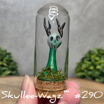 Glass dome with a skull faced creature and moss, held by a hand with a blurred green background.