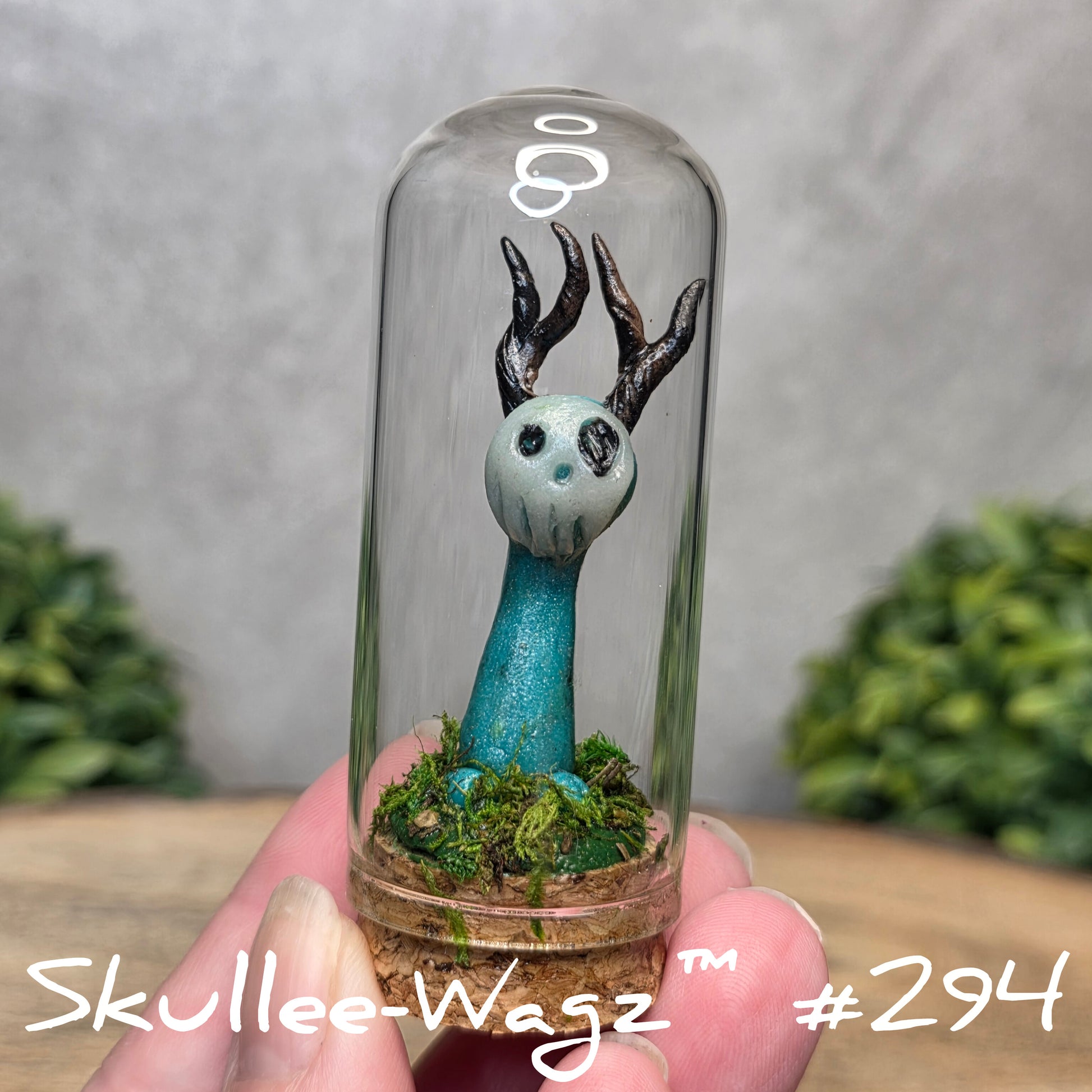 Glass dome with a skull faced creature and moss, held by a hand with a blurred green background.