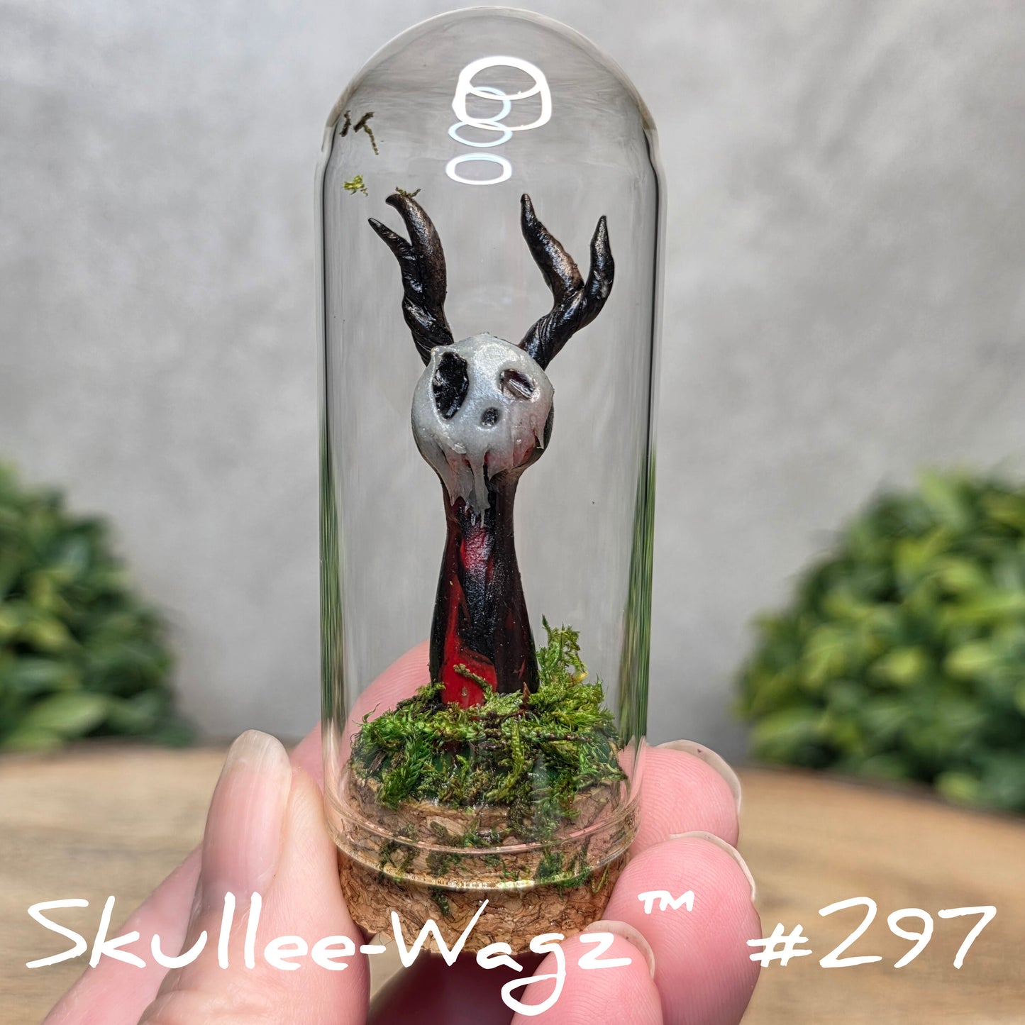 Glass dome with a skull faced creature and moss, held by a hand with a blurred green background.