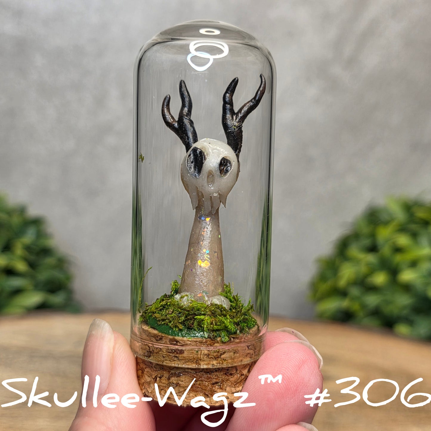 Glass dome with a skull faced creature and moss, held by a hand with a blurred green background.