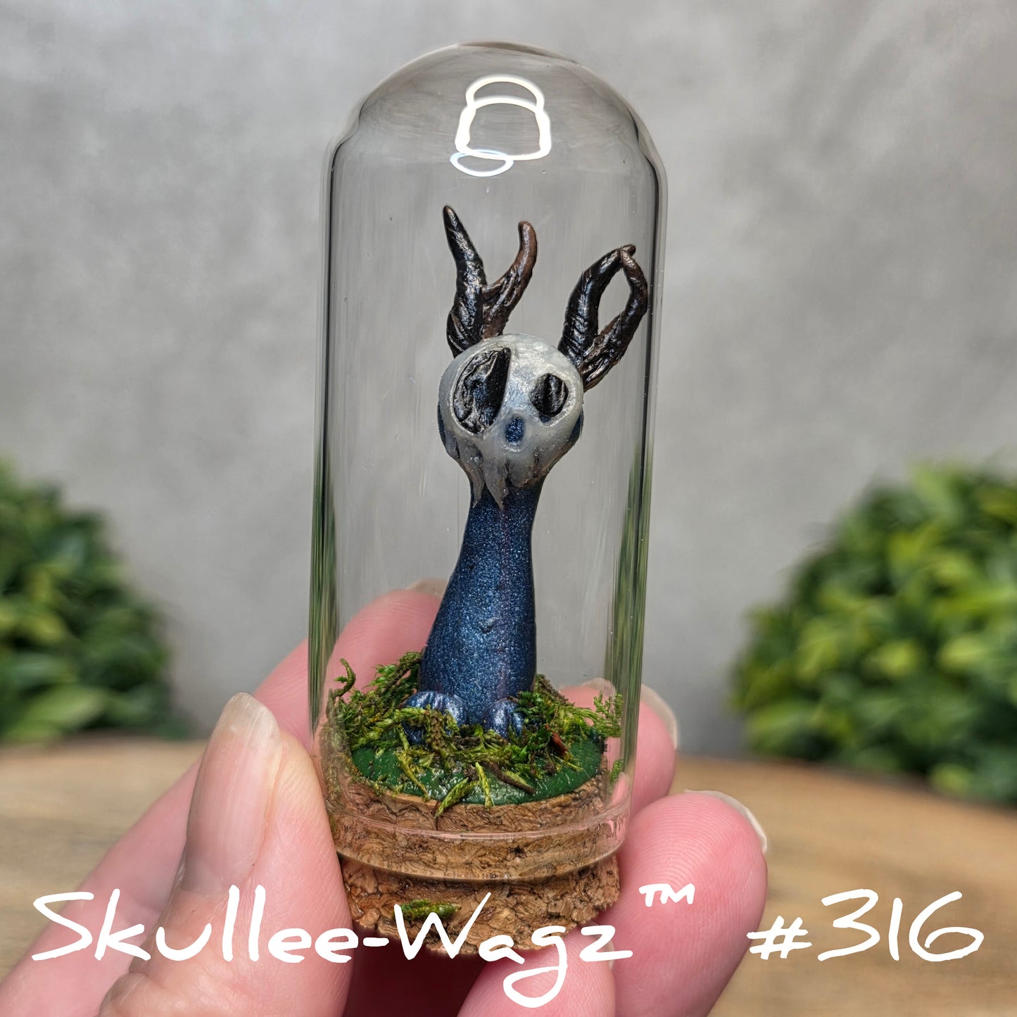 Glass dome with a skull faced creature and moss, held by a hand with a blurred green background.
