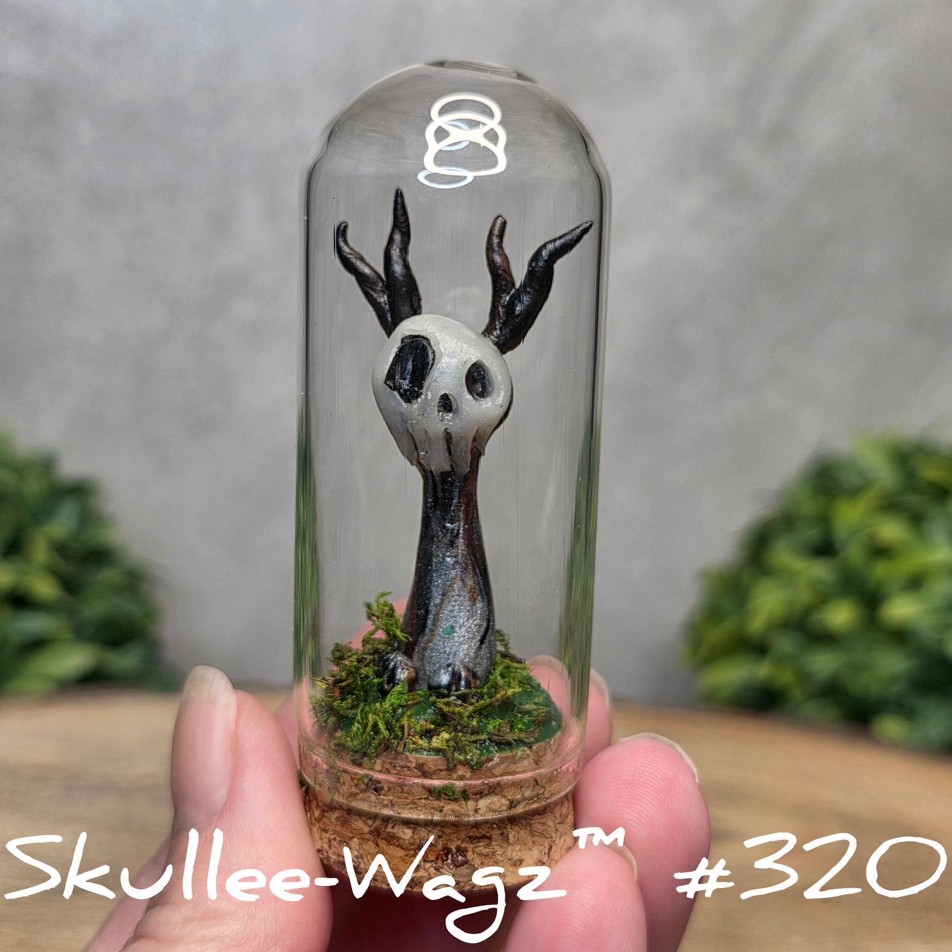 Glass dome with a skull faced creature and moss, held by a hand with a blurred green background.