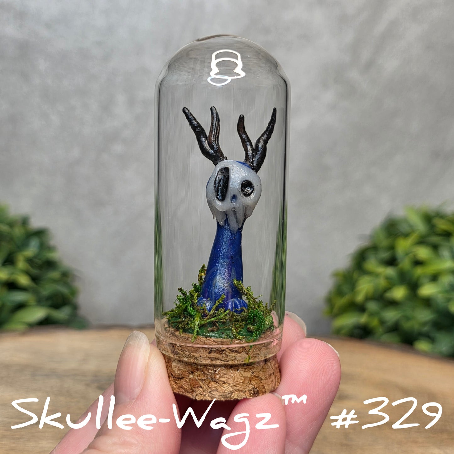 Glass dome with a skull faced creature and moss, held by a hand with a blurred green background.