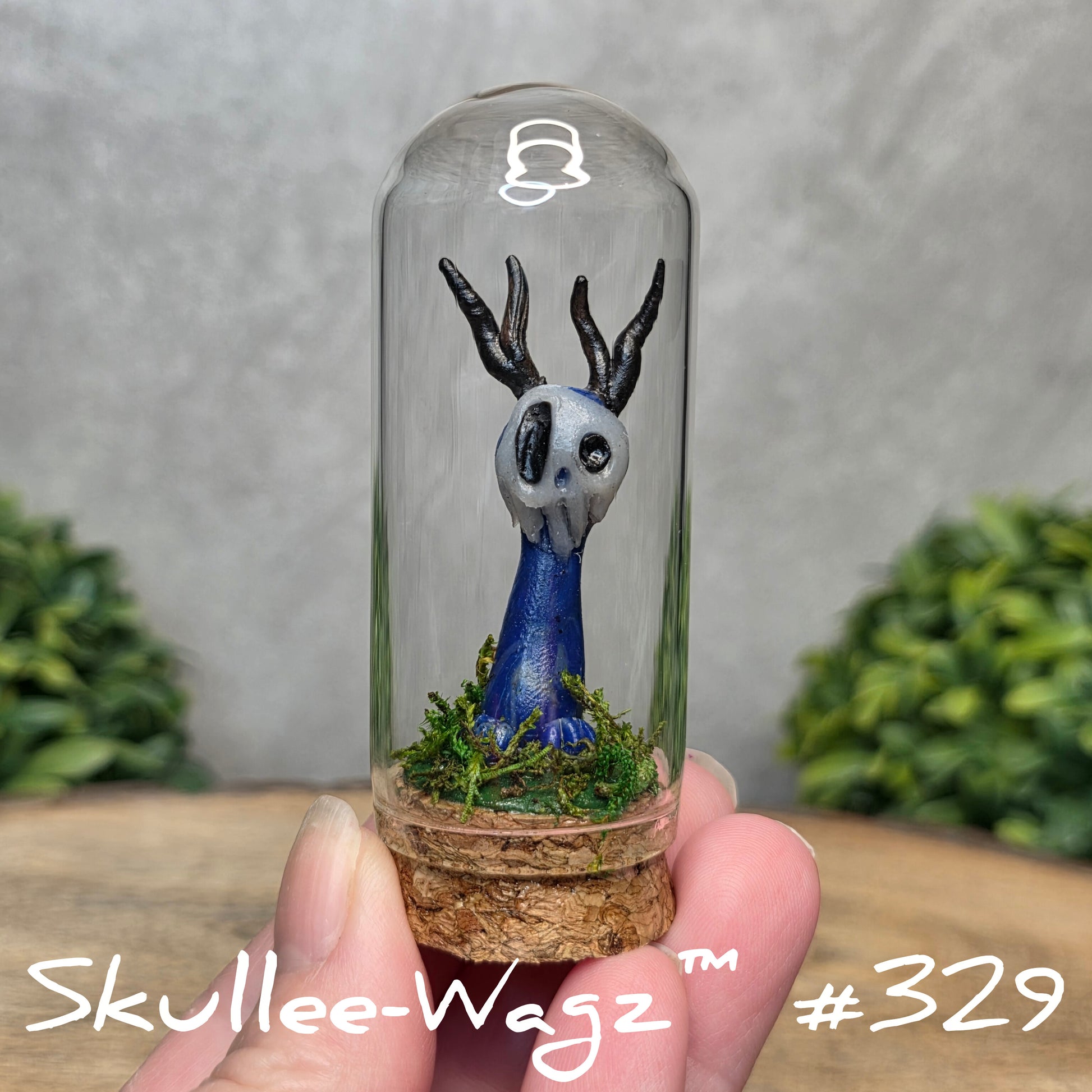 Glass dome with a skull faced creature and moss, held by a hand with a blurred green background.