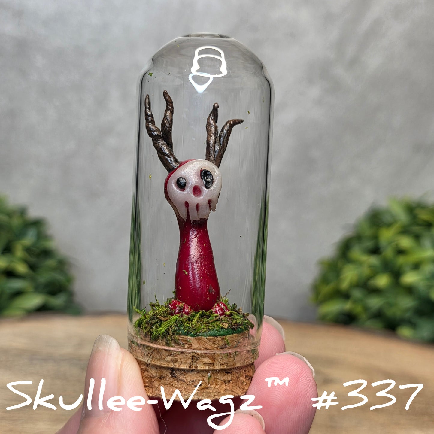 Glass dome with a skull faced creature and moss, held by a hand with a blurred green background.