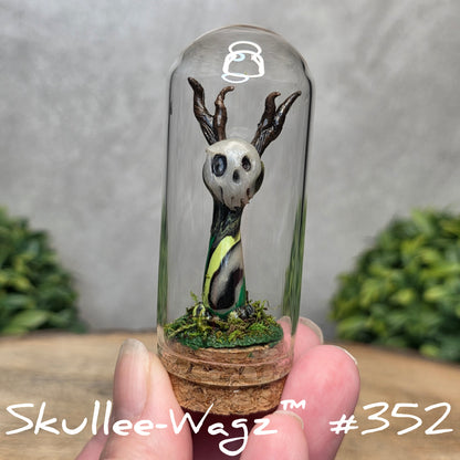 Glass dome with a skull faced creature and moss, held by a hand with a blurred green background.