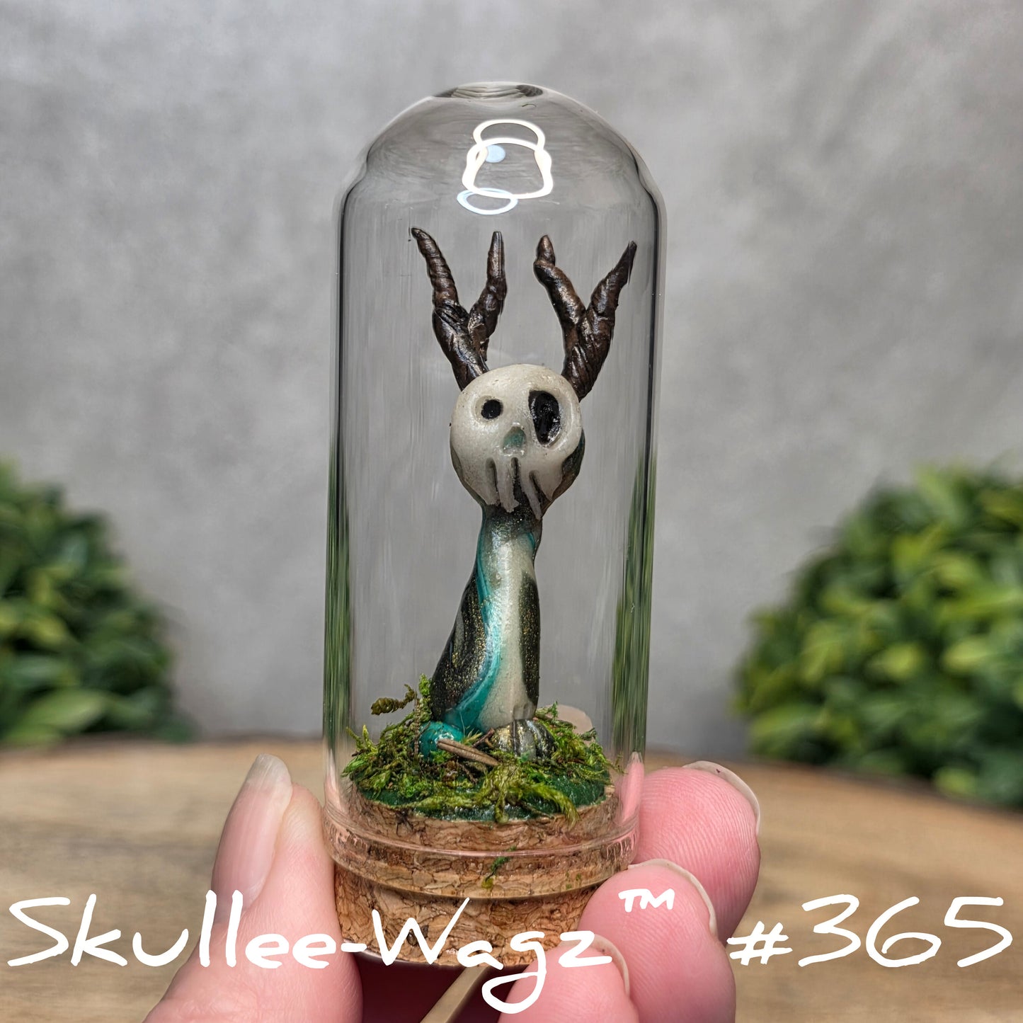 Glass dome with a skull faced creature and moss, held by a hand with a blurred green background.